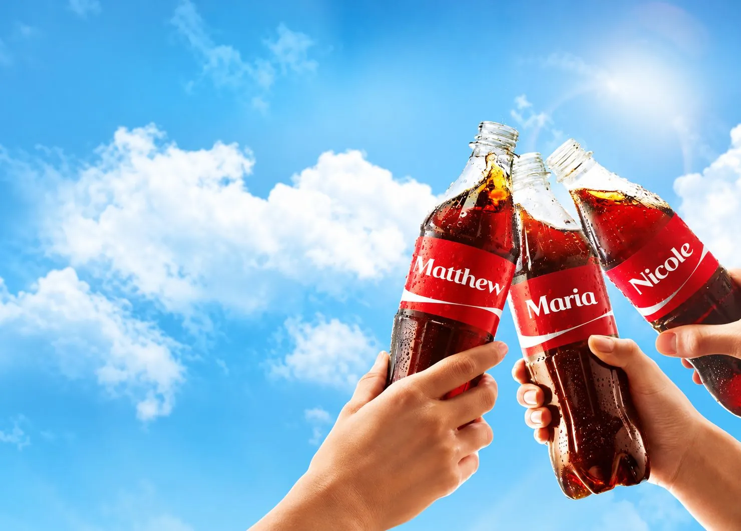 Three hands clink Coca-Cola bottles labelled Matthew, Maria and Nicole against a blue sky.