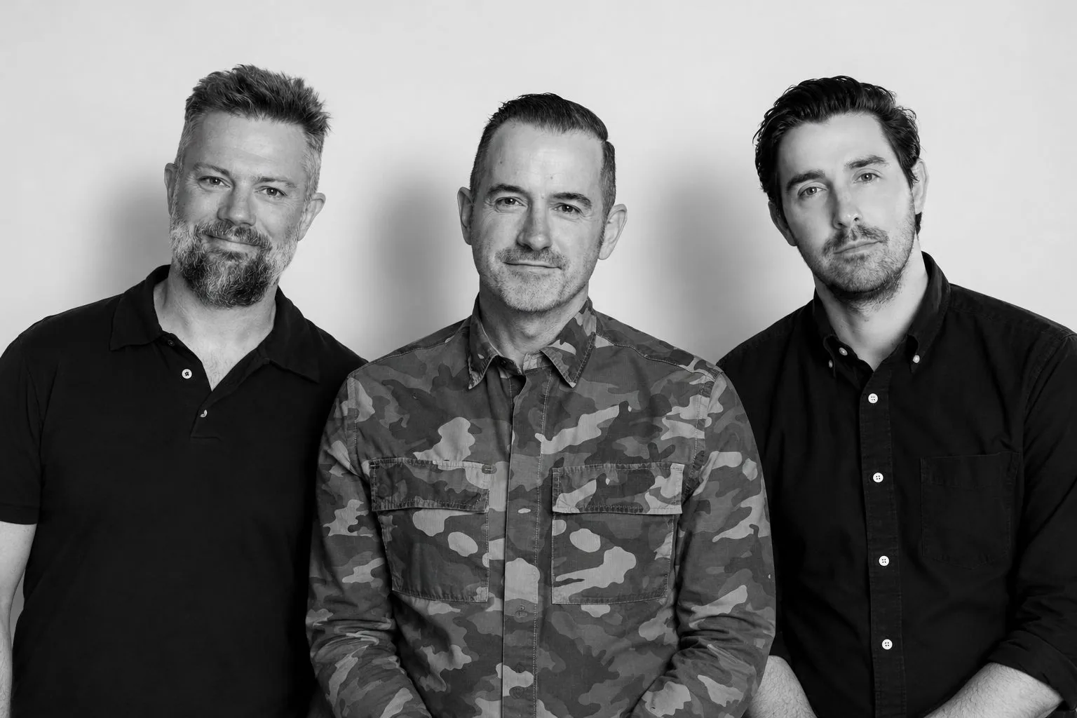 Toby Harrison, Boris Garelja and Andrew Hankin standing side by side against a pale wall, in black and white.