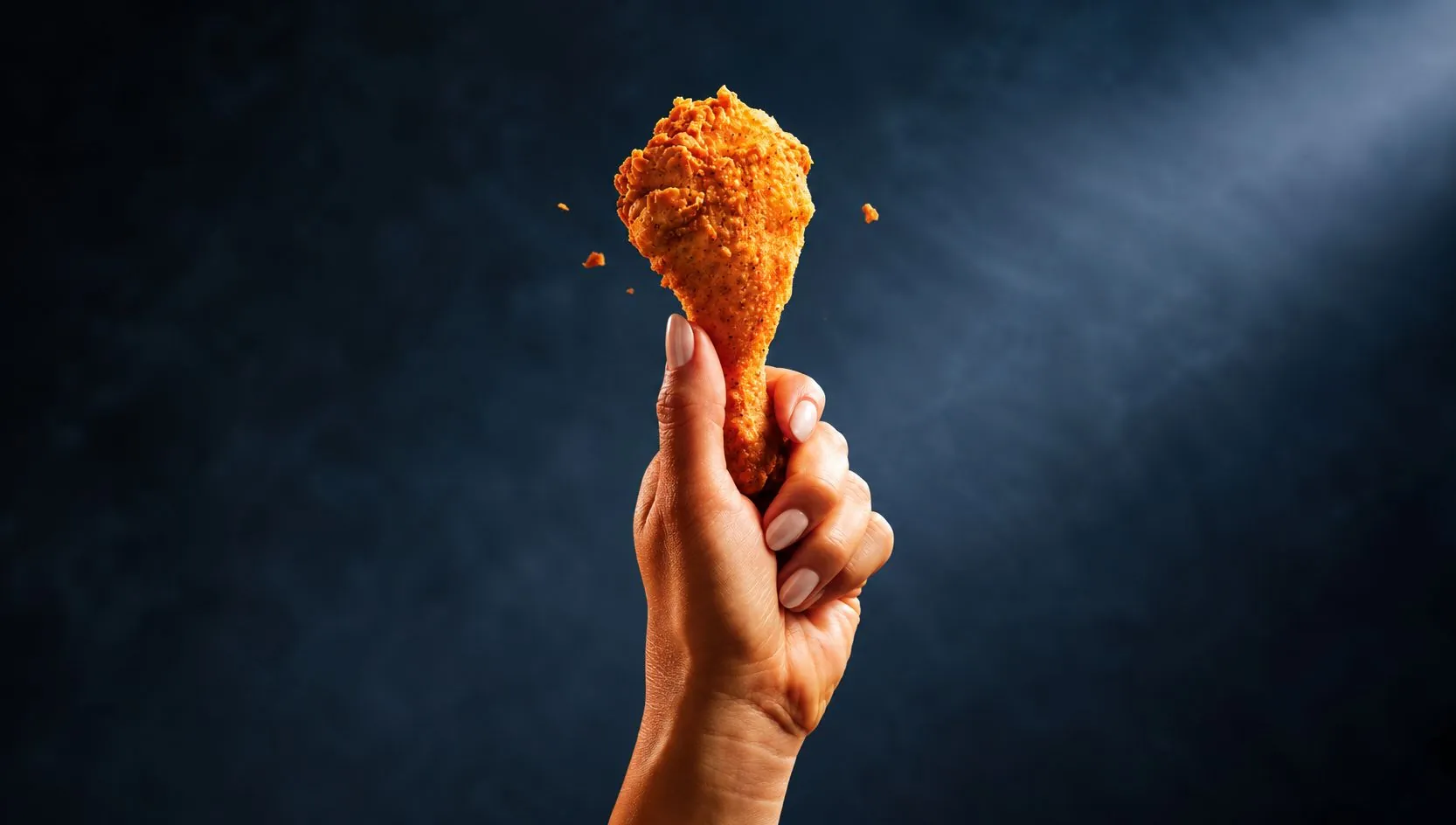 A hand holds up a piece of fried chicken against a dark blue background.