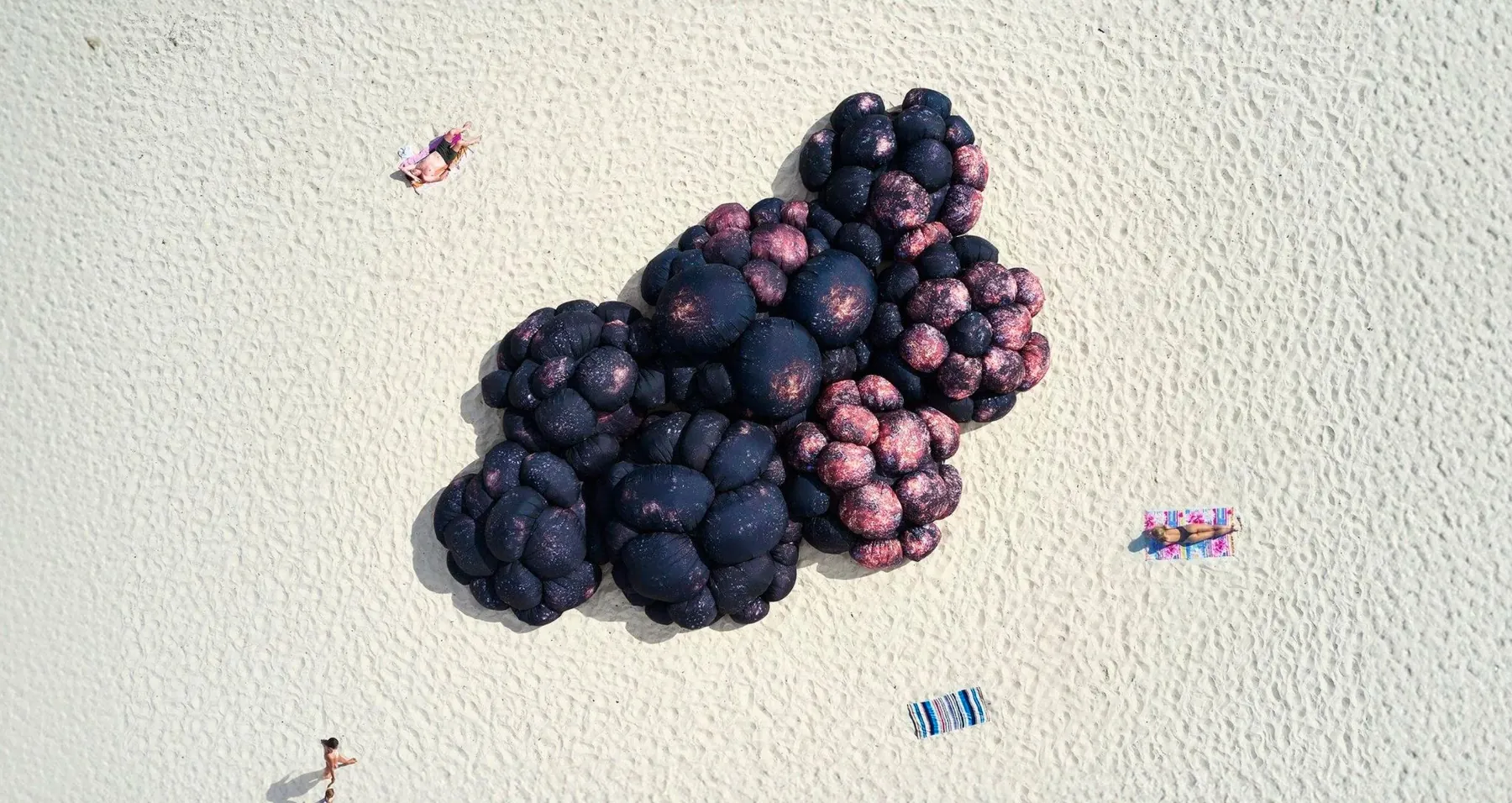 An aerial view of a giant dark, textured melanoma sculpture on a pale beach, with sunbathers either side.